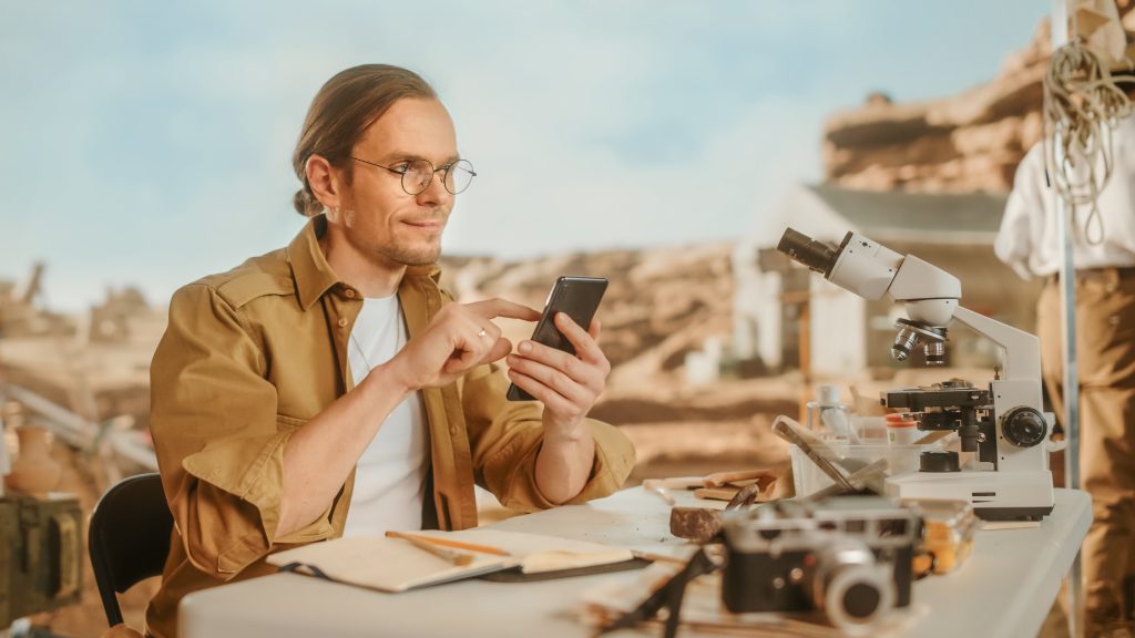 Archaeological Digging Site: Great Male Archaeologist Doing Research, Uses Smartphone to Share Discovery of Fossil Remains, Ancient Civilization Culture Artifacts on Internet Social Media